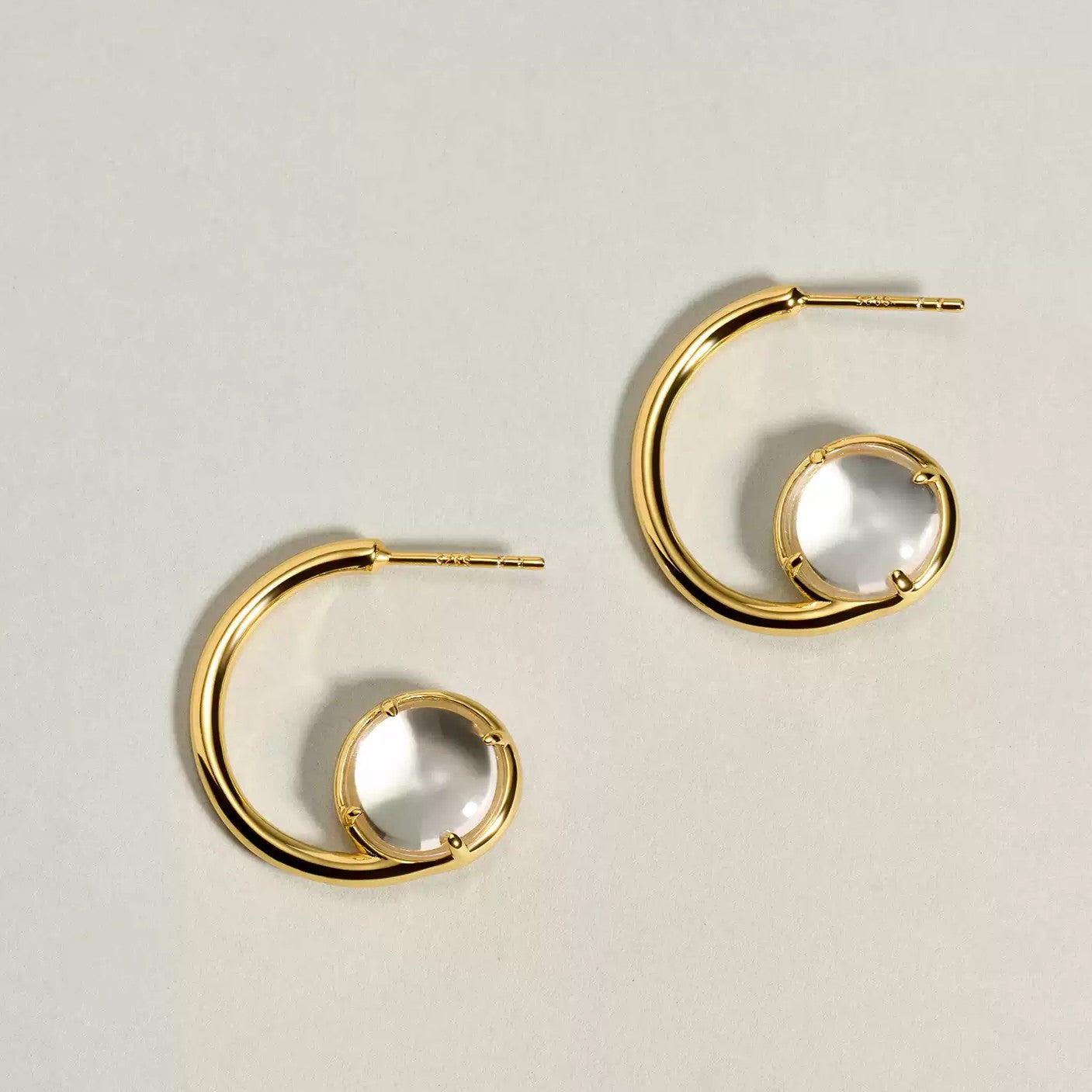 Pair of 18K gold plated 925 sterling silver G-shaped open swirl hoop earrings featuring natural round clear quartz cabochons in a four-prong setting on a light grey background